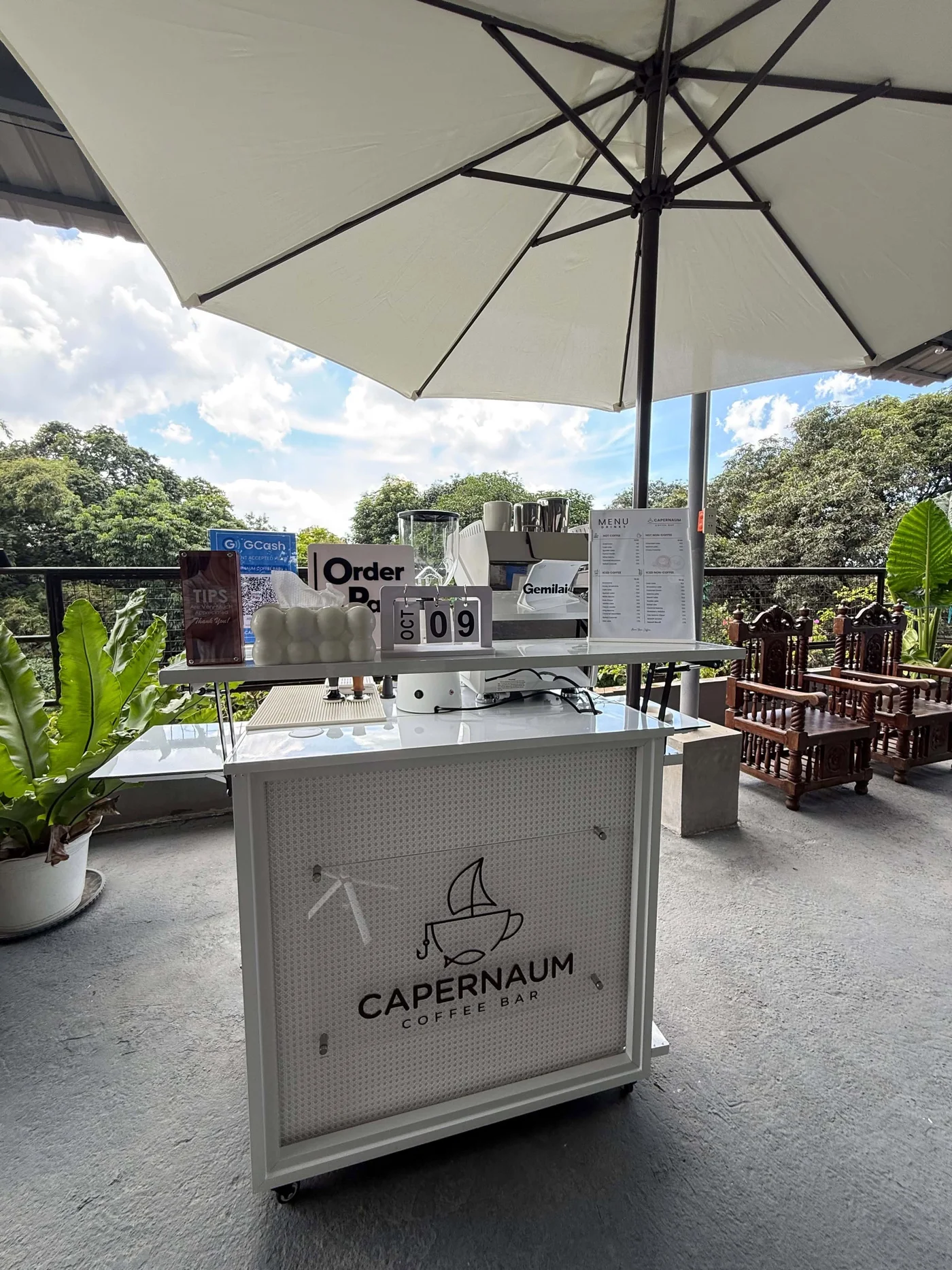 The Capernaum Coffee Bar mobile cart with its espresso machine and menu, set up under a cream umbrella.